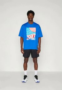 Kék rövid ujjú póló, nyomtatott kosárlabda grafikával, piros és fehér részletekkel; fekete rövidnadrággal és fehér zoknival párosítva.