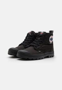 Stivali mid-top neri realizzati in materiale sintetico con suola in gomma. Presentano toppe con logo prominenti e lacci robusti. Supporto alla caviglia texturizzato.