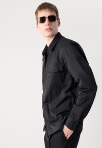 Young man wearing black zip-up jacket and sunglasses, posing with one hand in pocket against plain light background.
