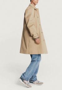 Manteau beige avec une coupe droite, orné de boutons et d'un col, associé à un jean bleu clair et des chaussures de sport multicolores.