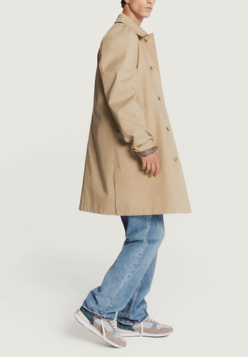 Manteau beige avec une coupe droite, orné de boutons et d'un col, associé à un jean bleu clair et des chaussures de sport multicolores.