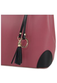 Roze handtas van nep leer met zwarte accenten, voorzien van een gouden ring en tassel detail aan één band, en een ronde zwarte basis.