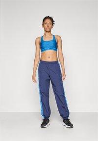 Sujeción deportiva azul con tejido texturizado y logo de Under Armour, combinada con pantalones de chándal azul marino con rayas laterales azules y cintura elástica.