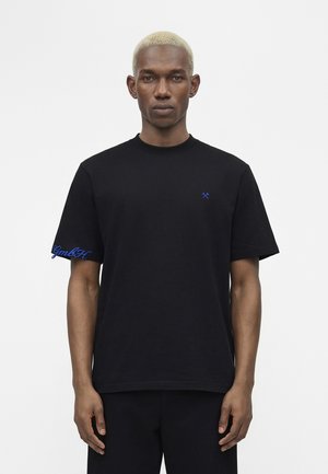 Homme aux cheveux blonds courts portant un t-shirt noir avec de petits marteaux bleus croisés sur la poitrine et un texte bleu sur la manche gauche.