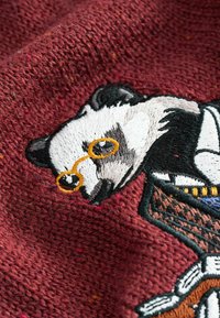 Czerwony dzianinowy materiał z haftowanym pandą w okularach, szczegółowo wykończony czarną, białą i żółtą nicią.