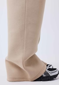 Pantalones deportivos beige con diseño de pierna ancha, hechos de material suave, combinados con zapatillas deportivas negras con detalles plateados y malla.