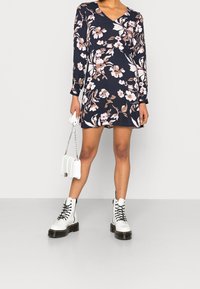 Vestito lungo a maniche lunghe floreale in blu navy con fiori rosa e bianchi, design con scollo a V, abbinato a stivali bianchi chunky e una piccola borsa bianca.