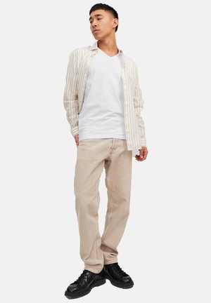 Jeune homme debout, les mains dans les poches, portant un pantalon beige, un T-shirt blanc à col en V, une chemise ouverte à rayures claires et des chaussures à lacets noires.