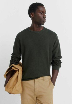 Maglietta a maniche lunghe a coste verde scuro abbinata a pantaloni beige chiaro. La maglietta presenta una scollatura arrotondata e maniche aderenti.