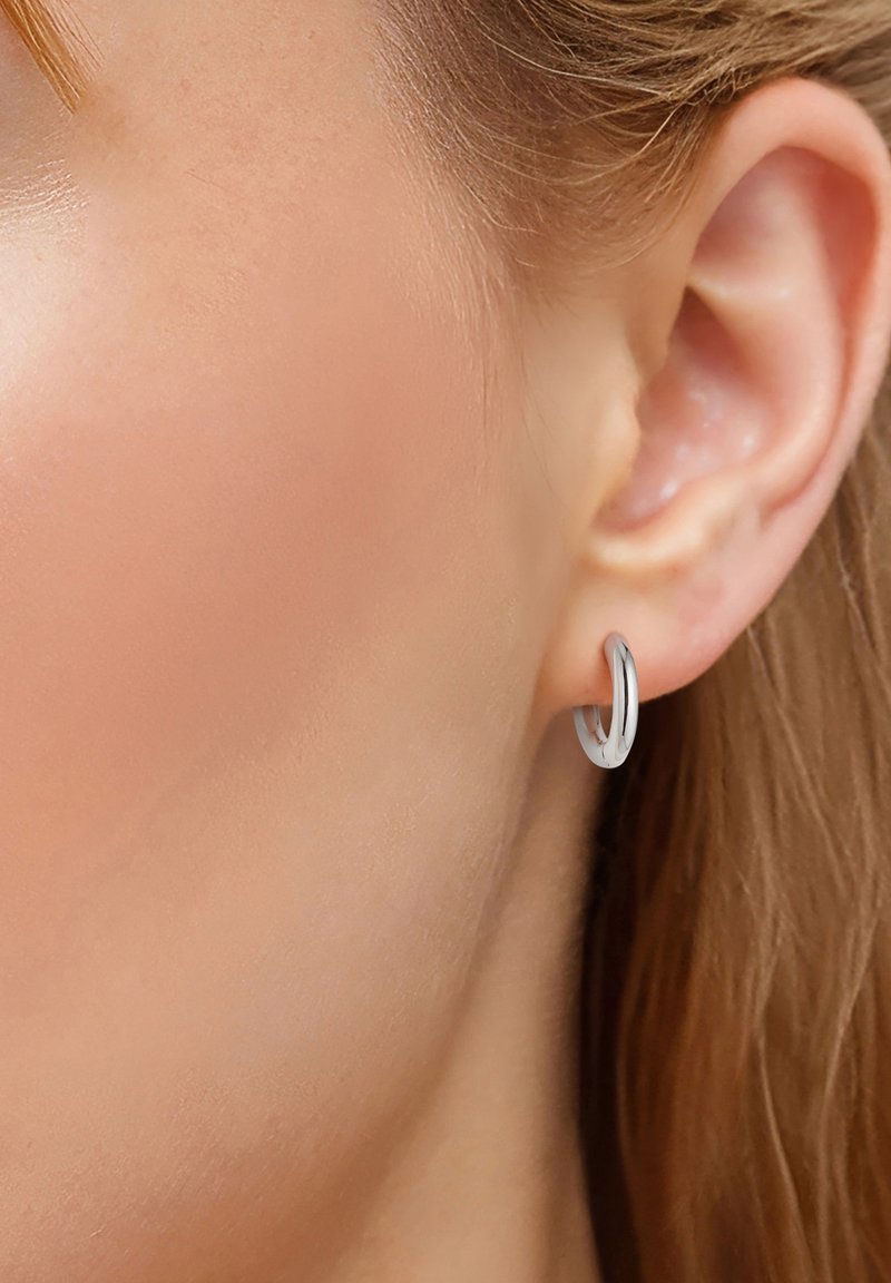Strieborný kruhový náušnice s lesklým povrchom, ktorý má štíhly, obručový dizajn s hladkou textúrou, zobrazené na uchu modelky.