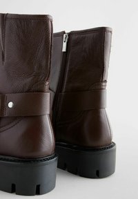 Botines de cuero marrón con un acabado texturizado, un cierre lateral con cremallera y una suela negra chunky. Cuenta con una correa decorativa y herrajes plateados.