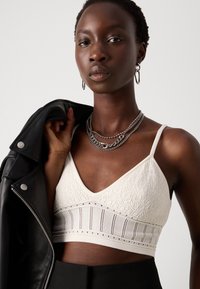 Weißes Textur-Bralette mit V-Ausschnitt, filigranen Mustern und verstellbaren Trägern, kombiniert mit einer schwarzen Lederjacke und geschichteten silbernen Ketten.