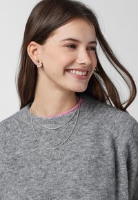 Collane di argento a strati con catene sottili e un maglione grigio a maglia texturizzata. Piccole orecchini di perle e metallo completano il look.