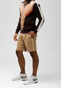 Miestenska farebne rozdelená športová bunda v hnedej, béžovej a červenej farbe so zipsom, v kombinácii s béžovými šortkami a bielymi teniskami s čiernymi akcentmi.