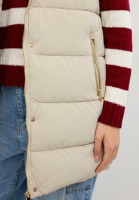Chaleco de plumas beige con un bolsillo con cremallera y herrajes dorados, con un diseño acolchado. Llevado sobre un suéter a rayas rojas y blancas.
