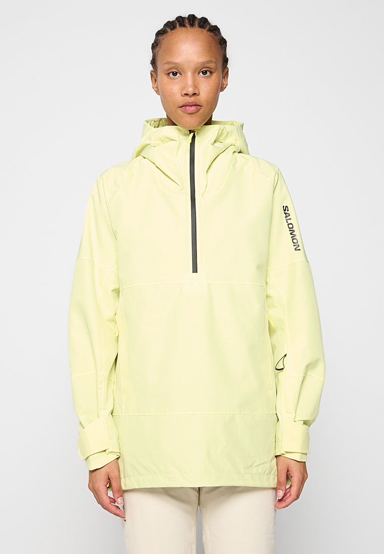 Mujer con trenza que lleva una chaqueta con capucha de color amarillo pálido, con una cremallera negra en el frente y el texto "Salomon" en la manga.