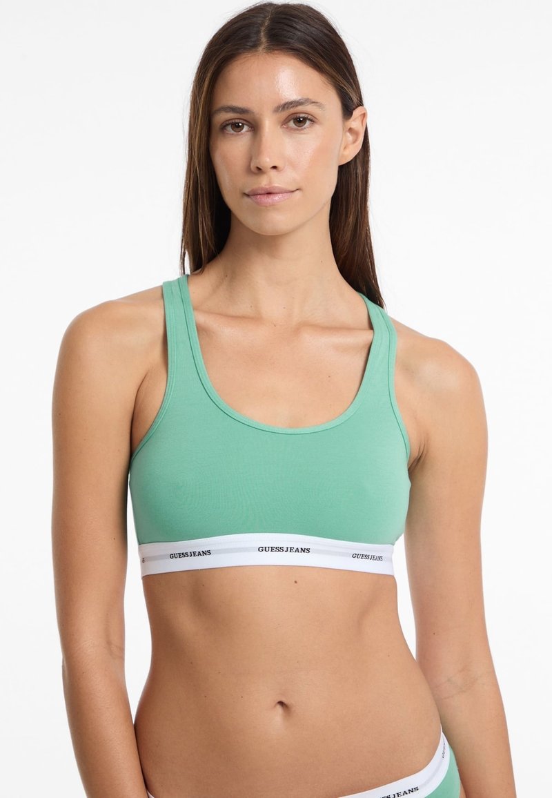 Groene sportbeha van zachte stof met een ronde halslijn en brede schouderbanden, voorzien van een witte elastische band waarop "GUESS JEANS" in zwart is gedrukt.