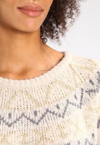 Stickad tröja med en krämfärgad bas, med grå och gyllene geometriska mönster. Ribbad halsringning och mjuk textur synlig.