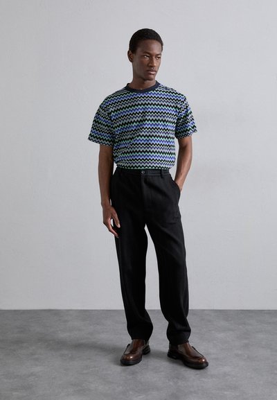 Lühikese varrukaga särk sinise ja rohelise zikk-zakk mustriga, koos mustade tekstureeritud pükstega ja tumeda pruuniga kingadega.