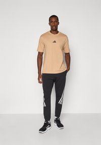 T-shirt beige clair avec logo noir Adidas, associé à un pantalon de jogging noir avec des rayures blanches sur les côtés, complété par des chaussures de sport noires.