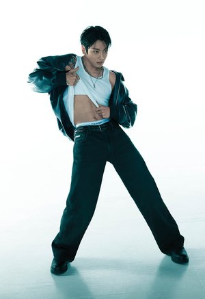 Muž v čiernej koženej bunde, bielom tielku a širokých čiernych nohaviciach dvíha tričko, odhaľujúc tetovania a pás Calvin Klein, pózujúc sebavedomo.