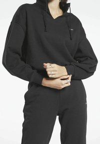 Osoba v černé mikině s kapucí a odpovídajících černých kalhotách, s rukama založenýma v pase, ozdobený řadou prstenů na prstech.