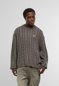 Férfi laza szürke kötött pulóverben és olajzöld nadrágban áll, az egyik kezét zsebében tartva, egyszerű háttér előtt.