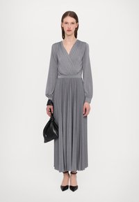 Robe plissée grise avec un décolleté enroulé plongeant, des manches longues et une taille cintrée. Associée à une pochette noire et des escarpins à bout pointu.
