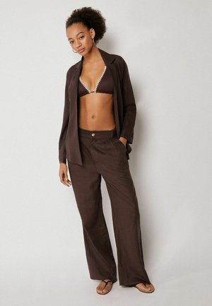 Žena se zvlněnými vlasy, oblečená v hnědém saku, kalhotách s širokými nohavicemi ve stejném odstínu, hnědém bikinovém topu s bílým lemem a černých žabkách, stojí před bílou zdí.