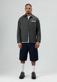 Zwart-grijs geruit overhemd met knopen en "FREEDOM" borduursel, gecombineerd met donkerblauwe denimshorts en sneakers, afgezet met witte sokken.