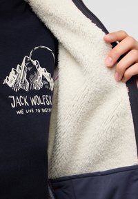 Giacca in pile navy con rivestimento interno crema, caratterizzata da una grafica di montagna e dal logo "Jack Wolfskin" in bianco. Tessuto morbido e testurizzato.