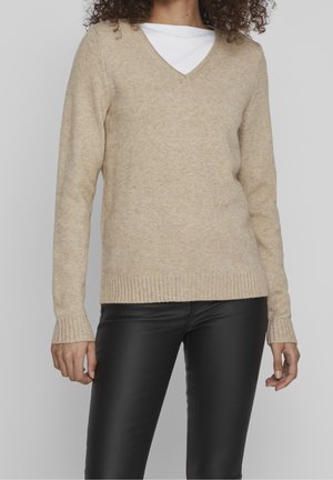 Pull beige en V en tissu doux et texturé, avec des poignets et un ourlet côtelés. Associé à un pantalon en cuir noir uni.