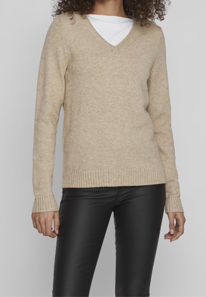 Beige V-halsad tröja i ett mjukt, strukturerat tyg, med ribbade ärmslut och nederkant. Matchad med enkla svarta läderbyxor.