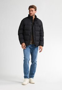 Uomo che indossa un giubbotto puffer nero sopra una camicia marrone, jeans blu e sneakers bianche, in piedi contro uno sfondo chiaro e semplice.