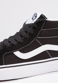 Baskets montantes noires avec une bande blanche, une semelle blanche, des lacets noirs et un logo visible sur la languette, sur un fond uni.