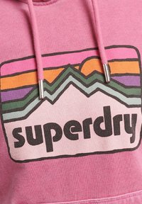 Felpa rosa con cappuccio caratterizzata da un design grafico con silhouette di montagne e un logo "superdry" in nero, con lacci e dettagli in argento.