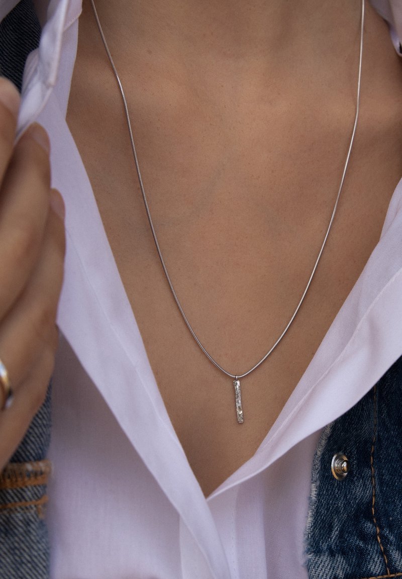 Zilveren ketting met een rechthoekige hanger met een gestructureerd oppervlak, hangend aan een fijne ketting, gedragen over een wit shirt.