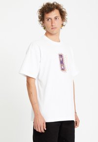Hvid bomulds t-shirt med korte ærmer, prydet med et farverigt grafisk design i midten. Modellen står foran en ensfarvet baggrund.