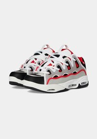 Un par de zapatillas deportivas blancas, negras, rojas y grises con suelas con amortiguación de aire y diseño de cordones sobre un fondo gris claro liso.