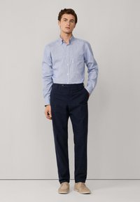 Giovane uomo in piedi, che indossa una camicia a righe blu chiaro con bottoni, pantaloni blu scuro e scarpe in camoscio beige, con una mano in tasca.