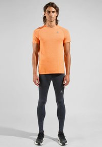 Elastische oranje T-shirt met korte mouwen van textiele stof, gecombineerd met nauwsluitende donkergrijze leggings. Beiden zijn geschikt voor sportkleding.