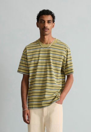 Man die een olijfgroen gestreept T-shirt met korte mouwen en lichtbeige broek draagt, staat met één hand in de zak tegen een effen grijze achtergrond.