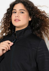 Svart zip-jacka med hög krage, har texturerade randdetaljer och en slät yta. Modellen håller i dragkedjans handtag mot en neutral bakgrund.