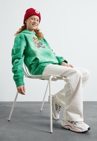 Green tie-dye hoodie with front graphic, white jeans, and gray sneakers. Sitting on a white chair against a light background.