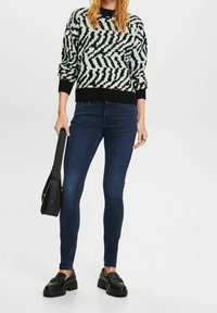 Suéter de patrón en blanco y negro con cuello redondo, combinado con jeans ajustados azul oscuro y mocasines negros. Lleva un bolso de mano negro.