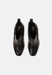 Un par de botas de cuero negro hasta el tobillo con paneles elásticos en los laterales y lengüetas para tirar, vistas desde arriba sobre un fondo claro.
