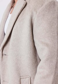 Cappotto grigio chiaro in lana con texture morbida, rever a lancia, due tasche frontali e vestibilità comoda, mostrato di profilo.