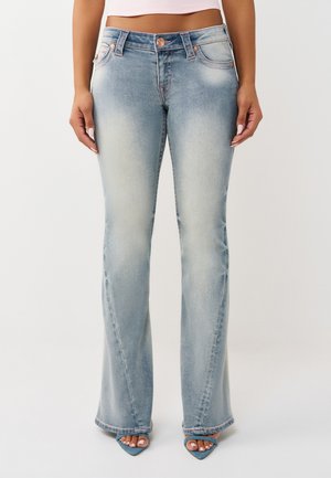 Femme portant un jean bootcut bleu clair délavé et des talons ouverts bleus, debout devant un fond blanc uni.