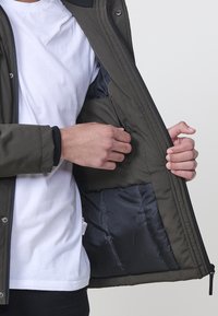 Olivovo zelená bunda s čiernou vnútornou podšívkou, so zipsom a zapínaním na cvoky. Obsahuje vnútornú kapsu na ľavej strane.