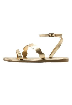 Sandale plate en métal doré avec bride à la cheville et bride ondulée au milieu du pied, sur une semelle fine, vue de profil sur fond blanc.